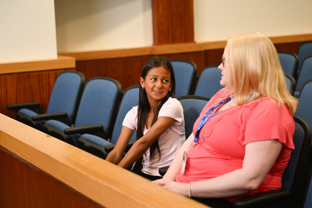 A young girl with long dark hair smiles at her CASA Volunteer, a blonde woman, as they sit in a courtroom.
